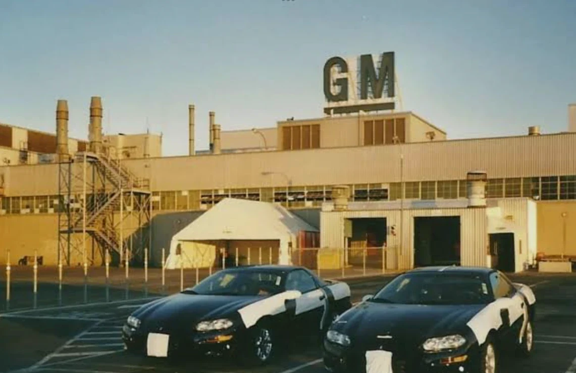 L'usine General Motors de Boisbriand, avec son enseigne GM sur le toit, et deux Camaro noires stationnées devant